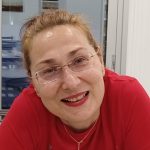A woman wearing a red tshirt and glasses smiles at the camera.